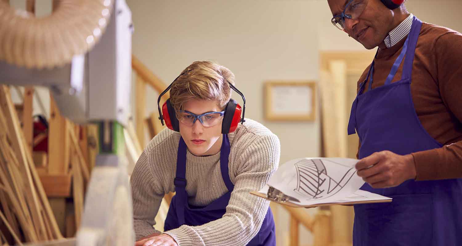 A young man with safety goggles on is watched by an instructor whilst he uses a table saw